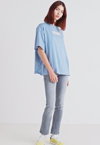 Lyseblå oversized t-shirt med et rektangulært "LEVI'S" logo, parret med falmede grå jeans og gule sneakers. Minimalistisk design, afslappet pasform.