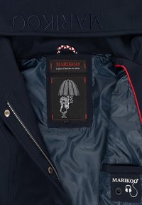 Marinblå jacka inre med slät tyg, röda accenter och en svart etikett med en karaktär under ett paraply. Dragkedja och knappar synliga.