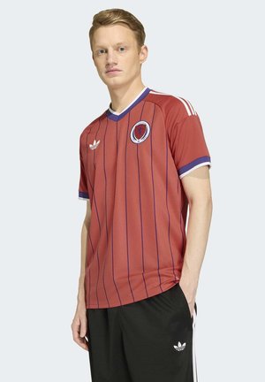 Junger Mann in einem roten Schottland-Fußballtrikot mit blauen Streifen und schwarzen Adidas-Hosen, der vor einem einfarbigen hellen Hintergrund steht.