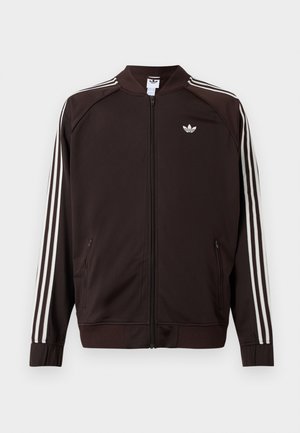 Σκούρο καφέ αθλητικό μπουφάν Adidas με λευκές τρεις ρίγες στα μανίκια, πλήρης φερμουάρ μπροστά και λευκό λογότυπο Adidas trefoil στο στήθος.