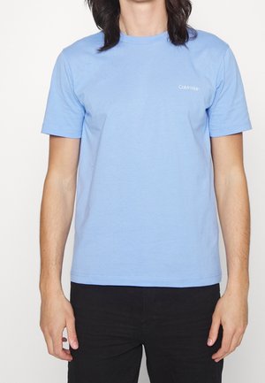 Adulte portant un t-shirt Calvin Klein bleu clair à manches courtes et un pantalon noir, debout avec les bras détendus le long du corps sur fond blanc.