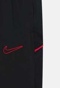 Uvećani prikaz crnog sportskog materijala s crvenim izvezenim Nike žigom i crvenom prugom na teksturiranoj crnoj tkanini.