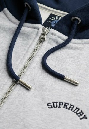 Grijze hoodie met ritssluiting en marineblauwe accenten, voorzien van een zilveren rits en marineblauwe trekkoorden met metalen uiteinden, geborduurd logo op de borst.