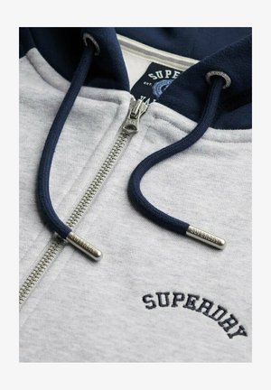 Grijze hoodie met ritssluiting en marineblauwe accenten, voorzien van een zilveren rits en marineblauwe trekkoorden met metalen uiteinden, geborduurd logo op de borst.