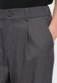 Σκούρο γκρι παντελόνια με πιέτες μπροστά, θηλιές ζώνης, κούμπωμα με κουμπί και χέρι μερικώς στην δεξιά τσέπη.