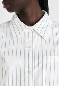 Primo piano di una persona che indossa una camicia bianca button-up con sottili strisce verticali nere e colletto a punta.