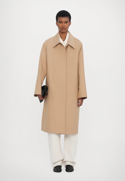 Beige wool coat with a wide collar, oversized fit, and long sleeves. Worn over white pants, the model holds a black clutch.