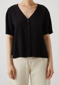 Blouse noire à manches courtes avec col en V, fermeture à boutons sur le devant et coupe décontractée, associée à un pantalon taille haute beige clair.