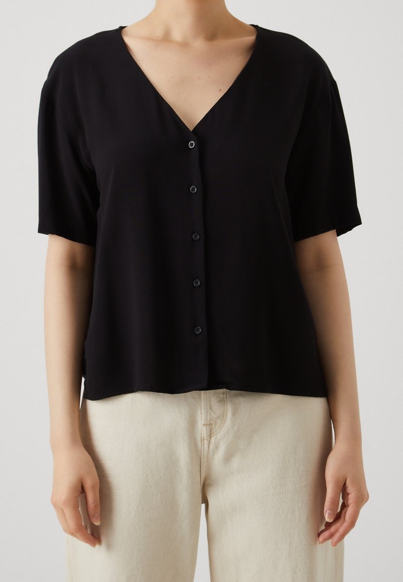 Blouse noire à manches courtes avec col en V, fermeture à boutons sur le devant et coupe décontractée, associée à un pantalon taille haute beige clair.