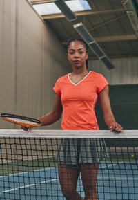 Camisola atlética laranja com acabamento branco, combinada com uma saia de ténis plissada cinza. A pessoa segura uma raquete de ténis preta e laranja junto a uma rede.