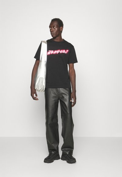 Black t-shirt with pink graphic "WHAT'S?" on the front, paired with black leather pants and a silver bag. Model stands against a white background.
