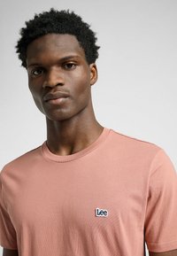 Jasnoróżowa bawełniana koszulka z okrągłym dekoltem i krótkimi rękawami, z małym, niebieskim haftowanym logo "Lee" na lewej piersi.