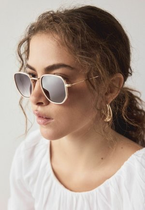 Ung kvinde med krøllet hår trukket tilbage, iført hvid indrammede solbriller, gyldne øreringe i hoop-stil og en hvid bluse med rynker, kigger til siden.