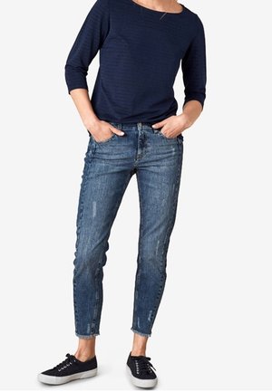 Vrouw draagt een marineblauw top met driekwartmouwen, versleten blauwe skinny jeans en zwarte lage sneakers met witte zolen.