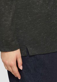 Donkergroene lange mouwen top van gestructureerde stof, met een zijsplit aan de onderkant. Hand rustend op de donkerdenim stof eronder.