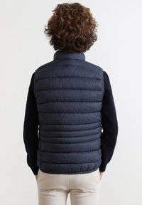 Marineblauer, gestepptes Pufferweste mit hohem Kragen, strukturiertem Stoff und horizontalen Nähten. Über einem dunklen Pullover getragen, maßgeschneiderte Passform.
