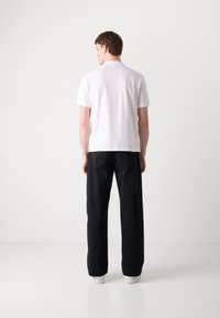 Hvit kortermet polo skjorte med flat krage, parret med mørke, løstsittende bukser med rette ben og hvite sneakers, sett fra bak. 