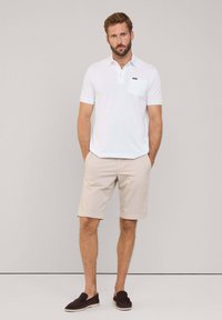 Biała koszulka polo z kieszonką na piersi, zestawiona z beżowymi szortami. Strój uzupełniają ciemnobrązowe loafersy. Prosty, casualowy design.