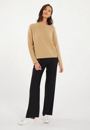 Pull en tricot beige avec un col rond et un petit logo sur la gauche. Associé à un pantalon large noir et des baskets blanches avec des accents rouges.