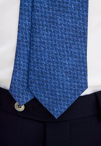 Sininen teksturoitu solmio, jossa on herringbone-kuvio. Solmion päät ovat terävät, ja se on aseteltu valkoisen paidan ja tummien housujen päälle.
