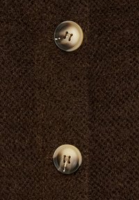 Brun texturerad tyg med en närbild av en metallknapp med fyra hål, som framhäver plaggets knapplistdetalj.