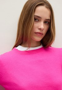 Suéter de color rosa brillante con cuello redondo y ribetes acanalados. Textura suave. El modelo tiene el cabello largo y está posicionado frente a un fondo neutro.