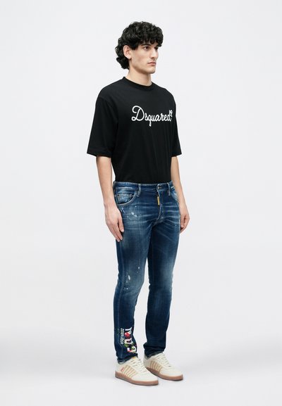 Μαύρο t-shirt με λευκό κείμενο "Dsquared2", συνδυασμένο με μπλε τζιν με φθορές και πολύχρωμα γραφικά. Το άτομο φοράει ανοιχτόχρωμα αθλητικά παπούτσια.