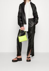 Svart faux läderjacka över en vit skjorta, kombinerad med svarta faux läderbyxor med slitsar, och en liten neongrön handväska. Svarta plattformssandaler.