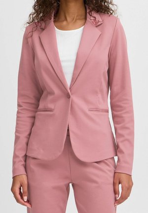 Femme portant un blazer rose poudré ajusté et un pantalon assorti, avec une chemise blanche côtelée en dessous, debout devant un fond uni.