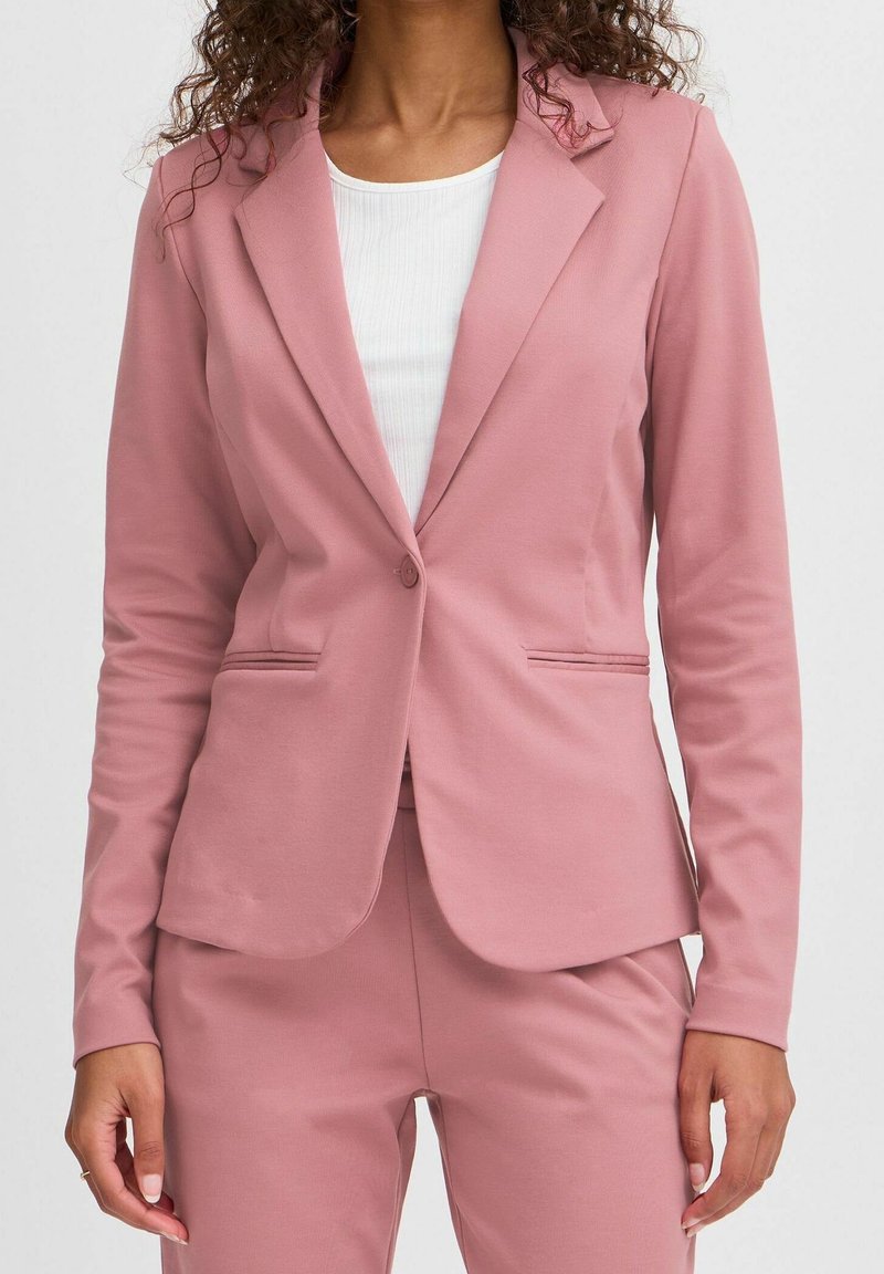 Femme portant un blazer rose poudré ajusté et un pantalon assorti, avec une chemise blanche côtelée en dessous, debout devant un fond uni.