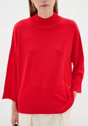 Femme portant un pull ample rouge vif à col rond haut et manches larges, associé à un pantalon clair sur fond uni.