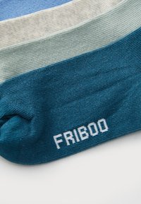 Stak af foldede strikkede sokker i blå, creme, mint og teal farver, med "FRIBOO" tekst på tåområdet af den teal sok.