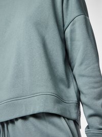 Nærbilde av overkroppen til en person som har på seg en løs, dempet grønn genser med lang erme og matchende bukser med synlige sømmer og en lomme.