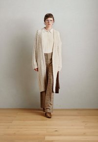Cardigan crema a maglia, blusa beige, pantaloni a quadri marroni e stivali con tacco marroni. Occhiali chiari, design strutturato, texture liscia.