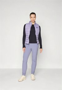 Ljuslila puffväst med vertikala quiltade sektioner, bärs över en svart långärmad topp, kombinerad med ljuslila avsmalnande byxor och beige skor.