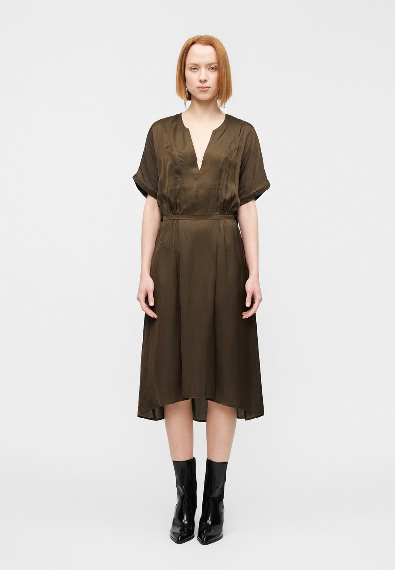 Φόρεμα ελιάς με κοντά μανίκια, βαθιά λαιμόκοψη σε σχήμα V, σφιγμένη μέση και ρέον στρίφωμα. Συνδυασμένο με μαύρα μποτάκια αστραγάλου. Ομαλή υφή υφάσματος.