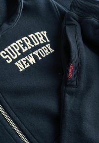 Sudadera con cremallera de color azul marino con la inscripción en crema "SUPERDRY NEW YORK" bordada; tejido texturizado, puños acanalados y un acento de etiqueta roja en la manga.