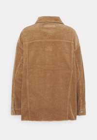 Veste en velours côtelé marron à manches longues, poignets boutonnés et détail de couture droite dans le dos sur fond uni.