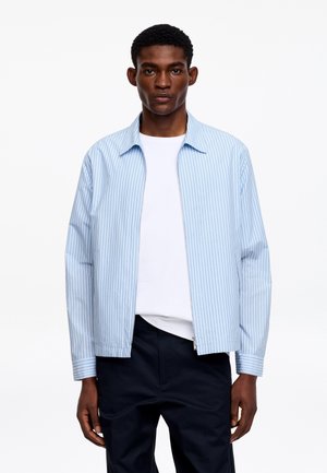 Młody mężczyzna w jasnoniebieskiej koszuli na zamek z pionowymi paskami, nałożonej na białą koszulkę, oraz w czarnych spodniach, stoi na jednolitym białym tle.