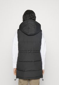 Svart vadderad väst med huva, med insydd midja, kontrasterande sömmar och justerbara snörningar vid fållen för en anpassad passform.