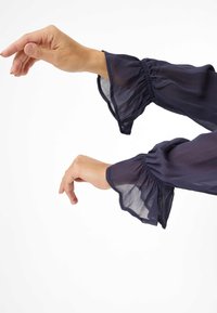 Maniche lunghe trasparenti di colore blu navy con polsini arricciati e orli svasati. Mani posizionate con grazia, in mostra la leggerezza e la fluidità del tessuto.