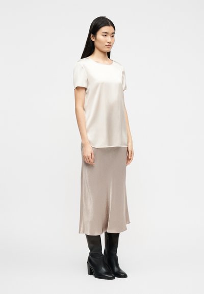 Γυναίκα με μακριά σκούρα μαλλιά, που φοράει μεταξωτό μπεζ μπλουζάκι με κοντά μανίκια, ταιριαστό midi φούστα και μαύρες ψηλές μπότες με τακούνι, στέκεται μπροστά σε λευκό φόντο.