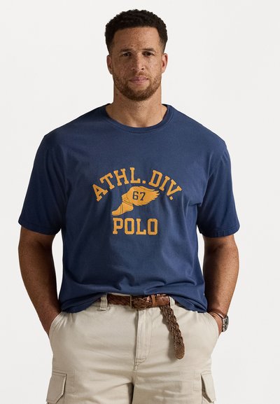 Navy blue cotton t-shirt with short sleeves, featuring orange graphic text "ATHL.DIV 67 POLO" and a wing design. Worn with beige cargo shorts.