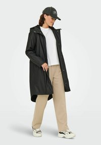 Frau trägt einen schwarzen kapuzenregenschutz, ein weißes Hemd, beige Hose, schwarz-weiße Sneakers und eine schwarze Mütze mit dem Text "Los Angeles".
