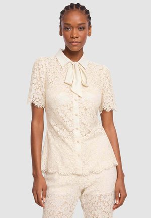 Donna che indossa una camicetta crema in pizzo a maniche corte con colletto a fiocco e pantaloni in pizzo abbinati, in piedi contro uno sfondo chiaro e uniforme.