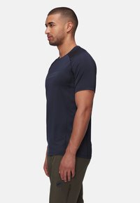 Marinblå sportt-shirt med kort ärm och rund halsringning. Har en subtil logotyp och en slät textur. Buren med olivgröna byxor.