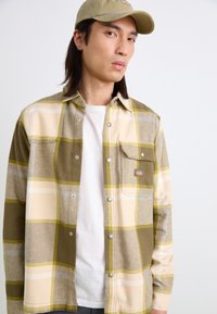 Chemise en flanelle à carreaux verts, beiges et jaunes ; comprend une poche et des boutons-pression. Portée par-dessus un t-shirt blanc, avec une casquette assortie.