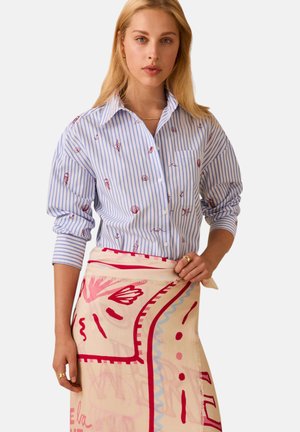 Donna bionda che indossa una camicia a righe blu e bianca con stampe rosse a tema marino e una gonna sciarpa crema con motivi astratti rosa e rossi.