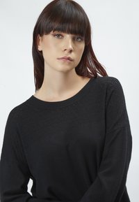 Schwarzer Pullover mit runder Halslinie, strukturiertem Strickmuster an der Oberseite, weiten Ärmeln und einem weichen, leicht lockeren Schnitt.