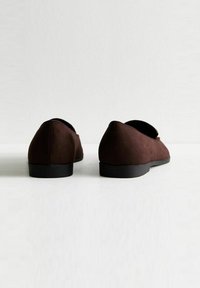 Zapatos de ante marrón tipo slip-on con puntera redondeada, diseño minimalista y suela de goma negra lisa. La vista trasera enfatiza la forma y la textura.